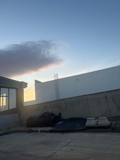 The image shows an outdoor urban scene in Pishtigovo, Bulgaria. The sky is blue with a large cloud. Part of a large, white, industrial building is visible. A metal ladder is attached to the side of the building. A gray concrete wall runs alongside the building. There are decorative planters and other objects. A modern building with glass windows is visible at the bottom of the frame. The lighting suggests it is either sunrise or sunset.
