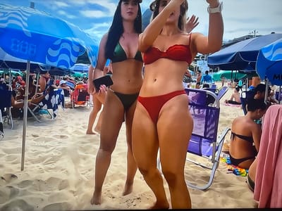 The image depicts a beach scene. In the foreground, two women are visible, both wearing bikinis. The woman on the left is wearing a green bikini and holding a black phone. The woman on the right is wearing a red bikini and appears to be taking a selfie. The beach is sandy, with other beachgoers and umbrellas visible in the background. The sky is cloudy. The location is identified as the city of Shakhovskaya, Russia.