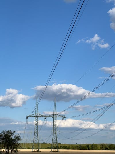 This media file shows a landscape scene with power lines stretching across a bright blue sky dotted with fluffy white and grey clouds. The power lines are supported by tall, green lattice pylons, two of which are prominently featured in the foreground and mid-ground. The pylons are connected by thick black cables that crisscross the sky.
In the background, a green, rolling landscape is visible with a line of trees and some distant structures that appear to be more power pylons. The sun is shining, suggesting it is daytime. The weather appears fair with scattered clouds, creating a dynamic sky. There are no people visible in the media file. The scene evokes a sense of vastness and industrial infrastructure within a natural setting. The location is specified as Újezd u Brna, Czechia. There is no visible text in the media file.