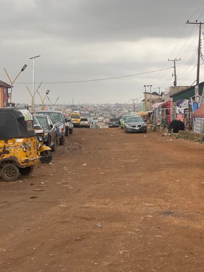 The image depicts a street in Ojodu, Nigeria, on a cloudy day. The road is unpaved and appears to be made of reddish-brown dirt. A line of parked vehicles, including a yellow "Keke Napep" (tricycle taxi), cars, and vans, are positioned on the left side of the street. On the left side of the street, there are buildings and commercial establishments lining the road. Electrical poles and wires are visible above the buildings. In the distance, a dense urban landscape stretches towards the horizon. No people are clearly visible in the image.