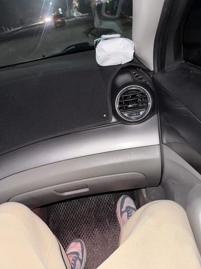 An interior view of a car reveals a gray dashboard with a circular air vent and a black fabric panel below. A white object is adhered to the windshield. The passenger seat, upholstered in a light, corduroy-like fabric, is partially visible. The floor mat is a dark, textured material, and two pink and blue sneakers are positioned on it, suggesting the presence of a passenger.