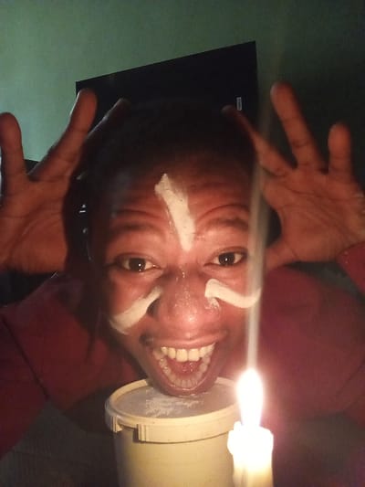 A person with white paint on their face is positioned behind a lit candle set atop a round, white container. The person's hands are raised above their head. The background includes a dark object that could be a television. The location is not discernible from the image.