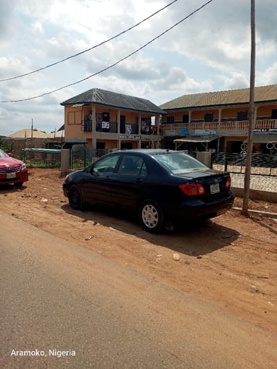 The scene depicts a black sedan, likely a Toyota Corolla, parked on the side of a paved road in Okinni, Nigeria, at coordinates latitude: 7.832, longitude: 4.51. The car is facing away from the viewer, and its license plate number is visible. The road is flanked by a dirt shoulder. The car is parked near a two-story commercial building with a sign that reads "MASTER BLA COFFEE SHOP" and another building further down the street. There are some electrical wires running across the scene. The sky is partly cloudy. The surrounding environment is characterized by a dry, dusty landscape. No people are visible in the scene.