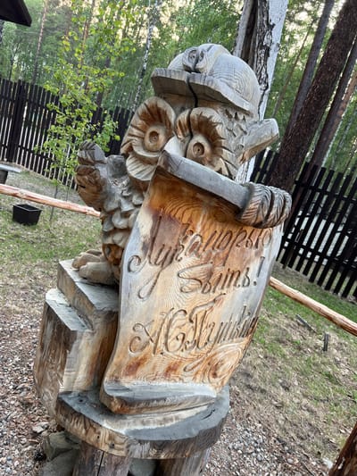 An outdoor shot features a wooden carving of an owl wearing a hat, holding a scroll with Russian text carved into it. The owl carving is on a pedestal made of logs. The scene is set amongst trees and a wooden fence, suggesting a park or forest location. The text on the scroll is partially legible, with the words "Добро пожаловать!" (Welcome!) and possibly an artist's signature. The location is identified as the Tal'tsy Museum of Architecture and Ethnography, near Irkutsk, Russia.