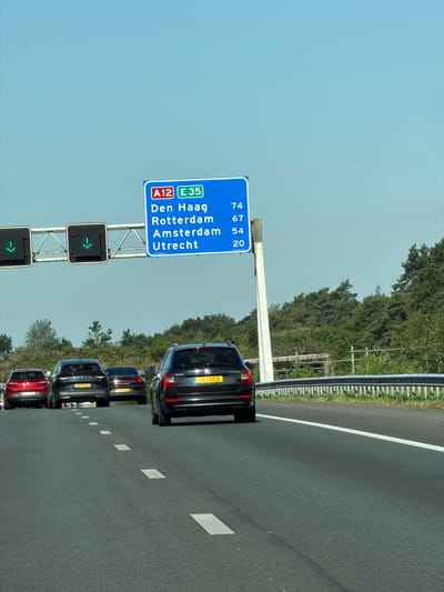 A highway overhead sign with blue background displays route information "A12" and "E 35" in red and green boxes respectively. Below the route information, a list of cities and their corresponding distances in kilometers is displayed: "Den Haag 74", "Rotterdam 67", "Amsterdam 54", and "Utrecht 20". The sign is supported by a white pole. To the left of the sign, two traffic lights with green downward arrows are visible. The scene is on a highway with several cars driving on it. The cars are a red sedan on the left, followed by two dark sedans, and a black wagon on the right. The background consists of lush green trees and a clear blue sky. The location is the A12 highway near Doorn, Netherlands.
