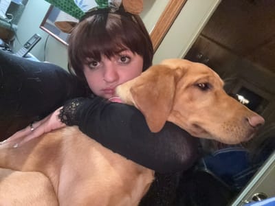 A woman with brown hair and bangs lies on her back, cradling a golden Labrador Retriever. She wears a black shirt with lace sleeves. The dog rests its head near her face. A piercing is visible in her nose. In the background is a wooden surface, an electrical outlet, and a framed picture. The angle of the photo is unusual.