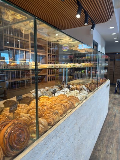 This is a forensic, matter-of-fact description of a bakery interior in Verin Ptghni, Armenia. The scene depicts a well-lit bakery with a focus on a refrigerated display case filled with various baked goods.
**Main Subjects:**
* **Baked Goods:** The primary subjects are an array of pastries and bread displayed within a glass-enclosed refrigerated case. These include numerous croissants, some sprinkled with sesame seeds and others with powdered sugar or almond slivers. There are also larger, swirl-shaped pastries, possibly cinnamon buns, prominently featured in the foreground. Several price tags with numbers such as "900", "1100", and "800" are visible among the pastries.
* **Display Case:** A large, multi-tiered glass display case dominates the foreground and mid-ground. It is constructed with clear glass panels and a light-colored, textured base that extends outwards. The interior shelves of the case are lined with paper or parchment.
* **Shelving Units:** In the background, wooden shelving units are visible, stocked with packaged goods, possibly snacks or other bakery items. Some of these packages have visible branding, including what appears to be a "Lay's" potato chip logo.
* **Ceiling and Lighting:** The ceiling features a slatted wooden design with integrated spotlights. Additional recessed circular lights are also present.
* **Floor:** The floor is covered with dark brown, wood-plank style flooring.
* **Staff Area:** In the far right background, a doorway is visible, marked with "STAFF ONLY" signage. A blue step stool is positioned near this doorway.
**Environment:**
The environment is clean and organized, suggesting a commercial establishment. The lighting is bright and functional, highlighting the products. The overall aesthetic is modern with natural wood elements.
**Actions/Interactions:**
There are no people visible in the image. The scene is static, focusing on the presentation of the baked goods.
**Objects:**
* Glass display case with refrigerated shelves
* Various pastries and bread (croissants, swirl buns, etc.)
* Price tags
* Wooden shelving units
* Packaged goods (chips, snacks)
* Wooden slatted ceiling
* Spotlights and recessed lighting
* Wood plank flooring
* "STAFF ONLY" sign
* Blue step stool
**Location Context (Verin Ptghni, Armenia):**
The presence of the bakery and the specific type of baked goods (croissants, buns) are consistent with a modern bakery offering both traditional and international items. The signage and overall presentation suggest a commercial retail space.