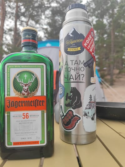 In a wooded area, possibly near Motornoe, Russia, a bottle of Jägermeister and a stainless steel thermos sit on a yellow wooden picnic table. The Jägermeister bottle is green with a prominent label featuring a stag's head and the brand name. The thermos is adorned with numerous stickers, including one with Russian text that translates to "I want to drink tea there" and another with "CCCP" in red lettering. In the background, a tent is visible, along with blurred trees, suggesting an outdoor setting. The lighting is overcast, indicating a cloudy day.