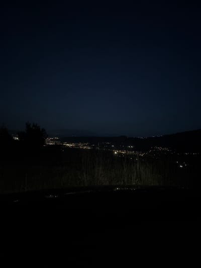The image captures a nighttime panoramic view of the city of Vrattsa, Bulgaria, seen from an elevated vantage point. The foreground is dominated by a dark, silhouetted landscape, likely a hillside covered in vegetation such as grass and trees, which are rendered as dark masses against the night sky. In the middle ground, the city lights of Vrattsa spread out across the valley. These lights appear as numerous small, bright points, suggesting a densely populated urban area. The lights are concentrated in a way that outlines the layout of the city and its streets. The sky above is a deep, dark blue, characteristic of a clear night with minimal light pollution from artificial sources other than the city itself. The distant horizon is visible, with the faint outline of hills or mountains further accentuating the topography of the region. The overall impression is one of a peaceful, illuminated city nestled within a dark, natural landscape.