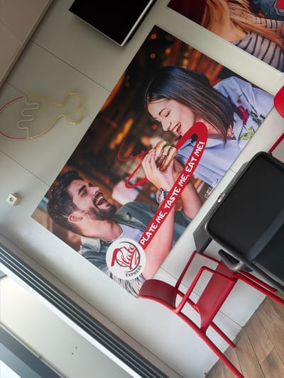 The image depicts an advertisement poster for “Hate Express” restaurant with a logo. The poster features a smiling couple, a man and a woman, sharing a hamburger and is attached to the ceiling or high up on a wall in a building. The text "Plate Me, Taste Me, Eat Me!" is printed around the hamburger. Part of a black serving tray or equipment is situated above the poster, as well as a red chair. Further, a neon art piece of a chicken is attached to the wall. The location is the city of Zlatna Panega, Bulgaria.
