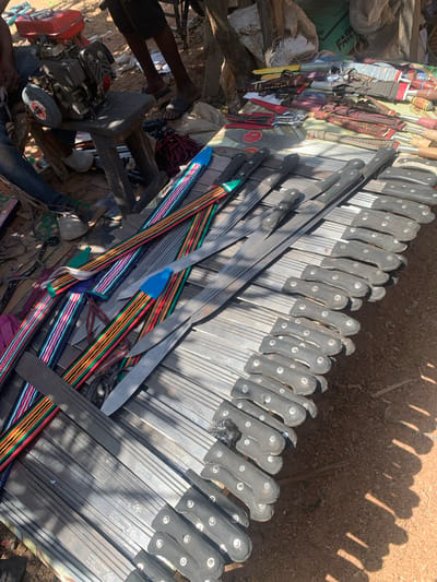 This media file shows a market stall in Nigeria, displaying a variety of items, predominantly knives and tools. The scene is set outdoors, with the ground appearing to be dirt and sand, suggesting a dry, possibly sunny environment, as indicated by the strong shadows.
The primary subjects are numerous knives, arranged in neat rows, with their blades facing towards the right side of the frame. These knives appear to be of various sizes and designs, some with black handles and others with more decorative, patterned handles featuring stripes of red, yellow, green, blue, and white. Some knives are sheathed in colorful, striped fabric. In the foreground and background, there are other objects, including what appears to be a small engine with a grinding wheel, some fabric, and bundles of string or rope. There are also hints of human presence, with legs visible at the bottom left of the frame, suggesting people are present at the market.
The activity taking place is likely the sale of these items at a market. The sheer number of knives suggests they are being offered for sale, possibly for agricultural use or as tools. The time of day appears to be daytime, given the strong sunlight and distinct shadows cast by the objects, indicating a clear day. The overall impression is one of a bustling outdoor market where tools and practical items are being traded.
Visible text on the knives includes "MADE IN ENGLAND," "NO 555W," "NASCENT CROOD," and "SARKON NOMA CROCODILE," suggesting the origin and possibly branding of some of the tools. The presence of "Project, Nigeria" in the location context further solidifies the setting as a market in Nigeria.