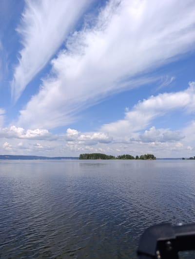 The image depicts a wide, calm body of water, likely a river or lake, with a distant tree-lined shoreline. The sky above is a vibrant blue, scattered with wispy white clouds, some of which are elongated and streaked, suggesting high-altitude winds. In the middle ground, several small, densely wooded islands break the expanse of water. The foreground shows the surface of the water with gentle ripples, and a dark, angular object, possibly part of a boat or a dock, is visible in the lower right corner, partially obstructing the view. The scene is serene and natural, evoking a sense of tranquility. The location is specified as Vekoshinka, Russia.