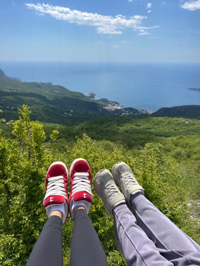 Two pairs of legs dangle over a landscape in Vojnići, Montenegro. The legs on the right are clad in gray athletic pants and gray sneakers. The legs on the left are clad in black athletic pants and red sneakers with white laces. Below the legs is a view of green trees, mountains, and water under a sky with scattered clouds.