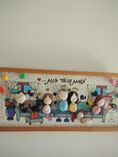 A framed artwork depicts a stylized family of five figures and a dog seated on a couch against a backdrop of a patterned wallpaper and a window. The figures, made of rounded objects, have simple facial features with dots for eyes and curved lines for smiles. The text above the figures reads "... Noah" and "New Year", written in Hebrew. The artwork is displayed on a wall in a home, likely in Pardes Hana-Karkur, Israel, suggested by the script. The overall scene is cheerful and family-oriented.