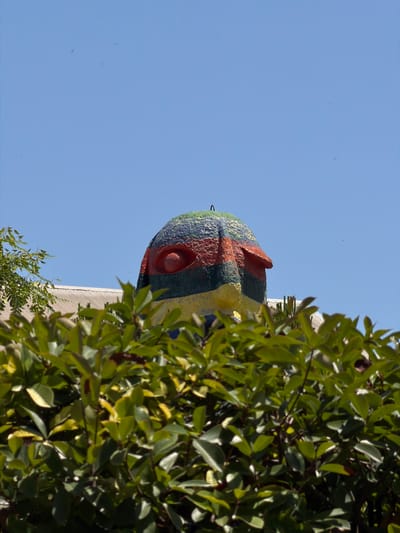 The image, taken in Castelló de la Plana, Spain, captures a close-up view of a colorful, mosaic-covered head sculpture positioned against a clear blue sky. The sculpture appears to be made of small tiles in various colors, including red, orange, yellow, green, and blue, and features a prominent red eye. A hook or loop is visible on the left side of the sculpture, suggesting it might be suspended or part of a larger structure.

The perspective is from below, looking upwards, with lush green foliage from a tree obscuring the right side of the frame and a portion of a building with a ribbed roof in the background. The lighting suggests it is daytime, likely a sunny day given the clear sky. The image seems to focus on the artistic details of the sculpture and its natural surroundings. There is no visible text in the image. The overall impression is one of vibrant art integrated into an outdoor environment.
