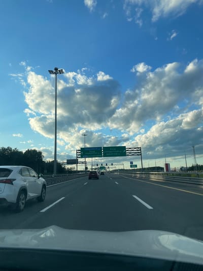 This is a photograph taken from the perspective of a driver on a highway in Pargolovo, Russia. The image captures a daytime scene with a bright blue sky dotted with fluffy white clouds. The highway is multi-lane and asphalt, with clear lane markings. On the left side of the highway, there is a white SUV. In the distance, several other vehicles are visible on the road.

Several large green highway signs are mounted on poles over the road. The signs display directional information and destinations in Russian. One sign indicates "Левашовское ш." (Levashovskoe Highway), "КАД" (Ring Road), "Приморское ш." (Primorskoe Highway), and "A181 Скандинавия" (A181 Scandinavia). Another sign points towards "КАД Комендантский пр-т" (Ring Road Komendantskiy Prospect) and "Выборгское ш." (Vyborgskoe Highway). Further down the road, a smaller blue sign with a white arrow and the number 12 is visible.

Tall streetlights are present along the highway, and on the right side of the road, there are trees and some industrial or commercial buildings in the far distance. The overall environment suggests a typical highway commute on the outskirts of a city.