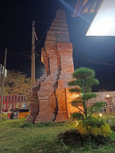 This is a nighttime outdoor photograph taken in Besuki, Indonesia. The main subject is a tall, stepped brick structure, possibly a monument or architectural feature, lit from below, casting a warm glow. In front of the structure, a small, well-maintained garden features a bonsai-like tree and other low-lying, textured plants, with yellow and dark green foliage.

To the left of the brick structure, a utility pole with multiple wires is visible against the dark night sky. Further to the left, a bright yellow car is parked, and a person is seated on the ground near it, possibly interacting with a blue object. Behind them, a row of red and grey rectangular structures with open fronts suggests a building or market stalls.

The scene is illuminated by artificial lights, indicating it is nighttime. The sky is dark and clear, with no visible weather phenomena. The presence of utility poles, a car, and the style of the background structures hint at an urban or semi-urban environment. The overall mood is calm and quiet, with the lighting creating a somewhat dramatic ambiance. No visible text is present in the image.