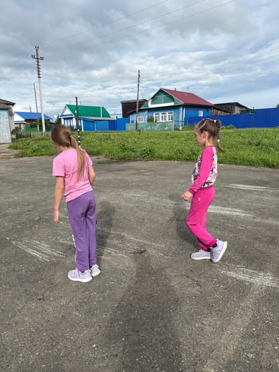 Two young girls are shown performing push-ups on a paved surface, likely a street or playground, in Большой Турыш, Russia. The girl in the foreground wears a pink t-shirt and purple pants, while the girl behind her is dressed in a pink floral long-sleeved shirt and pink pants. Both are wearing light purple sneakers. They are positioned in a push-up stance, with their bodies aligned and arms extended. The ground has white chalk markings.
The background features a grassy area and a row of colorful houses with blue and green roofs, typical of rural Russian architecture. Overhead power lines and utility poles are visible, indicating a residential or village setting. The sky is cloudy, suggesting overcast weather. The scene appears to be outdoors during daylight hours. The girls' focus and the athletic activity suggest a moment of physical exertion and perhaps playful competition. No visible text is present in the image.