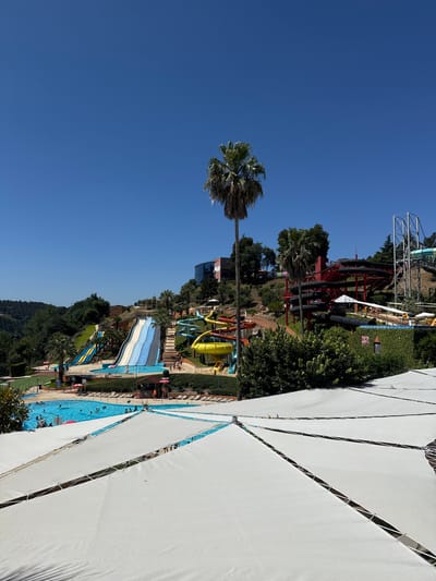 The image captures a sunny day at a water park. In the foreground, several white, triangular shade sails are stretched taut over a patio area. Beyond the sails, a large blue swimming pool is filled with people swimming and relaxing. In the mid-ground, a multi-colored water slide complex dominates the landscape. A tall, blue and white slide is prominent, with a yellow slide to its right and blue and yellow slides to its left. Further back, a steep, red structure with black coiled tubes represents another set of water slides. The park is set against a backdrop of lush green hills, with scattered palm trees and dense foliage. The sky above is a clear, vibrant blue. The location is identified as the Amarante Aqua Park in Amarante, Portugal.