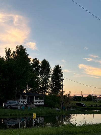 The image captures a serene sunset scene at a fishing spot in Kommunar, Russia. The sky is painted with hues of blue and orange, with wispy clouds catching the last rays of sunlight. A power line stretches across the upper left of the frame, silhouetted against the colorful sky.
The foreground is dominated by lush green grass and tall trees, creating a sense of natural abundance. In the middle ground, a small, white, open-sided structure with a pitched roof stands prominently, likely a pavilion or shelter for anglers. Several fishing rods are visible, suggesting an ongoing fishing activity. Two foldable chairs are set up in front of the structure, indicating people are present or have been recently. A dark-colored SUV is parked near the pavilion, its reflection shimmering in the calm water of the adjacent pond or river.
The water itself is a mirror, reflecting the trees, the structure, and the sky, adding to the tranquil atmosphere. The scene evokes a sense of peace and relaxation, typical of an evening spent outdoors. The time of day is clearly sunset, judging by the warm, soft light and the colors in the sky. The weather appears clear and calm, conducive to outdoor activities.
A yellow sign with black text is partially visible on the right side of the pavilion, but the text is not legible enough to decipher. The overall impression is one of a peaceful, natural setting with recreational activities taking place at dusk.