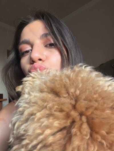 A close-up, selfie-style photograph captures a young woman with dark hair and makeup, puckering her lips as if for a kiss. Her face is partially obscured by a fluffy, light brown object, which appears to be the fur of a small dog. The background is dimly lit, suggesting an indoor setting. The image is focused on the woman's face and the furry object. There are no discernible features indicating the specific location within Aratashen, Armenia.