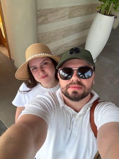 A man and a woman pose for a selfie. The man wears a green cap with a clover design, sunglasses, and a white shirt. He has a beard. The woman wears a straw hat adorned with pearls and a white t-shirt. They are outdoors, possibly near a building with light-colored tile or stone walls. A white vase with green plants is also visible.
