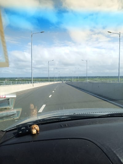 The image captures a forward-facing view from inside a vehicle, possibly driving or stopped on a multi-lane highway or bridge in Maputo, Mozambique. The asphalt road stretches into the distance with clear white dashed lane markings, flanked by concrete barriers and a continuous line of modern streetlights on both sides.

In the foreground, the vehicle's dashboard is visible. On the left side of the dash, there's a black electronic device, likely a dashcam, with "FUJIFILM" and "EV CABLE" faintly visible. Beside it, a small, light-colored, carved decorative object resembling a curled-up animal, possibly a turtle, rests on the dash. Reflections on the windshield show papers or documents on the left, with some blurry, partially legible text including "RIMERILA" and "RIA." A faint, distorted reflection is also visible on the windshield, superimposed over the road.

Beyond the road, a vast landscape extends to the horizon, featuring abundant green vegetation and what appears to be a body of water or an estuary in the distance. The sky above is partly cloudy with patches of bright blue. The scene is illuminated by natural daylight, indicating clear weather. The road ahead appears empty of other vehicles, suggesting open passage on this modern infrastructure.