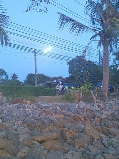 The image depicts a rocky shoreline in Baan Wok Tum, Thailand, under a twilight or dawn sky. In the foreground is a collection of rocks, varying in size and color. Beyond the rocks, a low hedge separates the shoreline from a row of buildings. A motorcycle is parked in front of one of the buildings. Power lines run across the sky above the buildings and palm trees. A street light illuminates the scene.