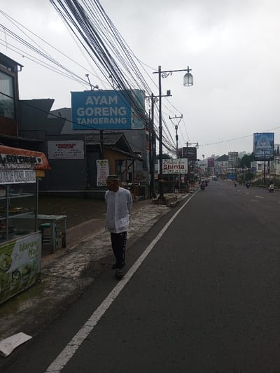 The image captures a street scene with overhead power lines. A sign advertises "Ayam Goreng Tangerang" (fried chicken Tangerang). Other signs are visible, including one for "Shinta." A man in a light-colored shirt and dark pants walks along the sidewalk. A food stall labeled "Warung Nasi" (rice stall) stands by the road. Motorbikes drive on the road. The sky is overcast.