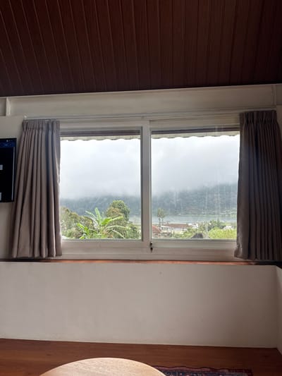 A double-paned window reveals a misty, mountainous landscape. The upper pane shows a foggy view with indistinct buildings. The lower pane features green foliage, including banana trees. Gray curtains frame the top and bottom of the window. The window is set between a wall with brown vertical paneling on the left and a plain white wall on the right. A dark electronic device is partially visible at the bottom center, below the lower curtain.