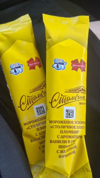 Two bright yellow ice cream wrappers are the main subjects in this image. The wrappers are oriented diagonally across the frame. The top wrapper is positioned slightly above and to the left of the bottom wrapper, creating a sense of depth. The wrappers appear to be made of a glossy material.
Both wrappers feature similar branding and text. Prominently displayed in large, stylized golden lettering is the word "Столичные," which translates to "Stolichnye" or "Capital City." Above this, in a red ribbon graphic, is the word "Коммунарка" (Kommunarka), indicating the brand. Below the large lettering, there is text in Russian, including "МОРОЖЕНОЕ ЭСКИМО" (ice cream eskimo) and "ПЛОМБИР" (plombir). There are also details about the flavor, such as "С АРОМАТОМ ВАНИЛИ" (with vanilla flavor), and descriptions of inclusions like "ШОКОЛАД" (chocolate) and "С ЖЕЛЕЙНОЙ НАЧИНКОЙ" (with jelly filling). A QR code is also visible on both wrappers. Small cartoon graphics of snowmen are present on the left side of each wrapper.
The background is dark and out of focus, suggesting the photo was taken in an indoor setting or in low light conditions, possibly with the wrappers resting on clothing or fabric. The lighting highlights the sheen of the wrappers. No people are visible. The overall impression is of consumer products presented for viewing.