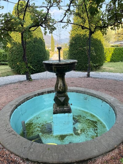 The image shows an outdoor fountain in a garden setting. The fountain is made of stone and has a tiered design, with water spouting from the top. The basin of the fountain is painted turquoise and contains some green algae. In the background, there are manicured green hedges, trees, and a gravel path. The scene appears to be in a temperate climate, possibly during the daytime, with natural lighting. The overall impression is one of a well-maintained garden with a central water feature. There are no people or animals visible in the image. The context of Balerna, Switzerland, suggests a European garden style.