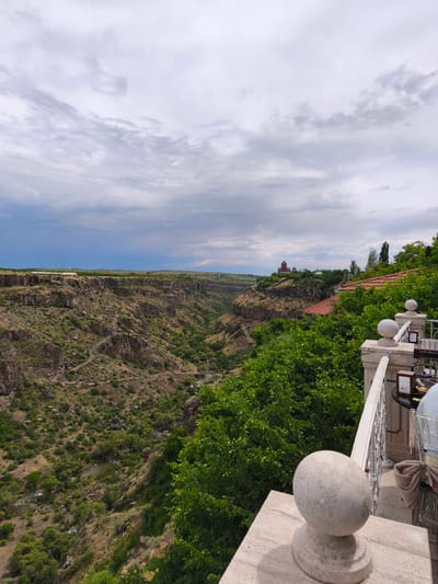 The image captures a scenic overlook in Ohanavan, Armenia, featuring a deep gorge and a distant monastery. In the foreground, a white railing and a decorative stone orb adorn a balcony, suggesting a vantage point for a restaurant or hotel. Lush green trees partially obscure the view. Beyond the railing, the gorge carved by a river stretches into the distance, its steep, rocky sides dotted with sparse vegetation. A winding path is visible on the gorge's slope. In the far distance, perched on the edge of the plateau, a multi-domed monastery with red-tiled roofs stands against a backdrop of rolling hills and a dramatic, cloudy sky. A hint of snow-capped mountains can be seen on the horizon. The lighting suggests an overcast day, with diffused light casting soft shadows. No people are clearly visible, but the presence of a table and chairs on the balcony indicates human activity.