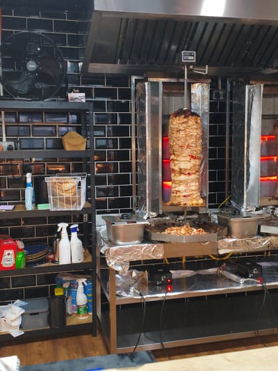 A commercial kitchen scene in Wychavon, United Kingdom, features a large vertical rotisserie with a substantial amount of seasoned meat slowly turning, illuminated by red heating elements. The rotisserie is housed within a stainless steel and foil-covered unit, with shaved meat collecting in a tray below. To the left, shelves are stocked with various cleaning supplies, including spray bottles and a red Heinz ketchup container, as well as a basket of what appears to be bread or flatbread. A black industrial fan is mounted on the wall above the shelves. The kitchen walls are covered in dark, glossy rectangular tiles. The overall environment suggests a food preparation area, likely for a restaurant or takeaway establishment, with an emphasis on hygiene and food safety indicated by the cleaning products.