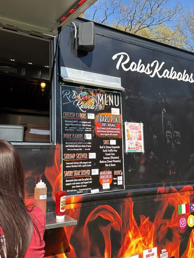 Here's a forensic description of the image:
**Scene:** The image depicts a food truck named "Rob's Kabobs" parked outdoors, likely in Rochester, United States based on the flag next to it. The truck is black with a flame design on the lower portion of the side visible.
**Subjects/People:** A person with long, dark hair and wearing a red shirt is visible in the foreground, obscuring the lower left portion of the frame. They appear to be a customer.
**Objects:**
* A food truck with a visible menu board.
* The menu board lists items such as Chicken Kabobs, Pork Kabobs, Shrimp Skewer, Savory Steak Skewer, Jibaro Plate, sides (Beef & Cheese Empanada, Garlic Truffle Fries, French Fries, Tostones, Maduros, Rice & Beans) , and drinks (Water, Soda).
* A "Sweet Specials" sign advertising desserts.
* Bottles of condiments (likely hot sauce and ketchup) on a small shelf attached to the truck.
* Flags: USA, Italy, and Puerto Rico
* Stickers: Instagram and Snapchat logos
**Environment:** The truck is situated in an outdoor setting. Bare trees are visible in the background, suggesting it might be early spring or late fall. The light suggests it's daytime.
**Actions/Interactions:** A person is apparently at the window to place or receive an order.
**Essential details:**
* The food truck's primary offering seems to be kabobs and other Puerto Rican dishes.
* There are visible "GAS SHUT-OFF" and "NO SMOKING" signs on the truck.