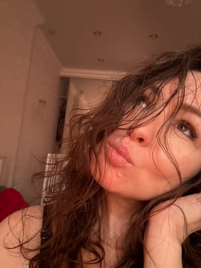 A close-up selfie shows a woman with dark, wet-looking hair framing her face. Her head is tilted, and she looks directly at the camera with brown/hazel eyes. Her lips are slightly pursed in a "kissy" expression, and a small blemish is visible on her right cheek. Part of her arm or hand is raised near her head. She wears a gold-colored chain or necklace. In the background, behind her, a light-colored wall with several circular indentations, suggestive of jacuzzi jets, is visible. Below, a reflective surface, possibly water or wet tiles, is present. The indoor lighting is warm and somewhat dim. A dark garment or strap is seen near her shoulder, and a piece of red fabric, possibly a towel, is visible at the bottom right. No text is visible in the image.