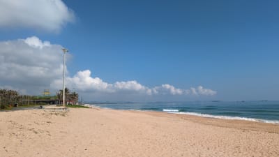 The image depicts a coastal scene in Thành phố Tuy Hòa, Vietnam, under a clear blue sky with scattered white cumulus clouds. The foreground and midground consist of a wide, light tan sandy beach with minor scattered debris. No individuals are visible on the beach. To the left, a paved road runs parallel to the coastline, separated from the sand by a low concrete structure. A tall, gray metal lamppost, fitted with multiple light fixtures, stands prominently beside this road. Behind the road, a low-rise building, partially obscured by dense palm trees and green foliage, features a yellow illuminated sign reading "SANDSBAY." Additional palm trees extend further along the coastal edge. In the midground, the turquoise ocean exhibits gentle waves breaking near the shore. Several dark-hulled vessels, likely fishing boats, are observable on the distant horizon. A low, darker landmass is also visible on the horizon towards the right side of the frame. The distant coastline features indistinct hills or mountains and faint indications of development. A white beach umbrella is partially visible on the far right edge of the land area.