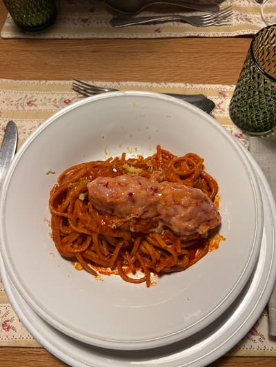 A plate of spaghetti with red sauce and two large, pale pink meatballs is centered on a white plate with a double-rimmed edge. The plate rests on a floral patterned placemat on a wooden surface. To the left are silverware (forks and a spoon) and a textured green glass. A knife lies at the bottom of the image near the plate's edge. The scene appears to be a meal setting. The location in the city of Gondarém, Portugal is not discernible from this single image.
