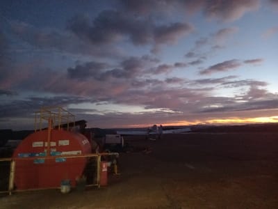 This image depicts a scene at dawn or dusk in Santa Juana, Chile. In the foreground, a large, horizontal, red industrial storage tank with yellow support scaffolding on top and yellow protective fencing around its base is situated on unpaved ground. Several white labels are affixed to the tank's side, and a red fire extinguisher is visible mounted to the fence structure. To the right of the tank, in the midground, a small, fixed-wing aircraft, likely an airplane, is parked on an open, unpaved area. The aircraft's fuselage and wings are partially visible, with its tail section extending toward the far right. No individuals are present. The background features a low horizon with silhouetted hills or terrain, beneath a sky displaying a gradient of colors from dark blue and purple at the zenith, transitioning through pinks and oranges, to bright yellow at the horizon, with scattered clouds.