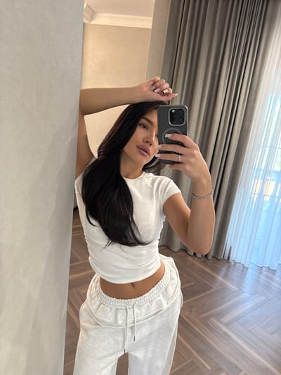 A woman is taking a selfie with a phone in front of a mirror. She is wearing a white t-shirt and grey sweatpants. The background includes a herringbone-patterned floor, a grey curtain, and a white window covering. The location is not explicitly stated as being in Дубровка, Russia, and there is no evidence in the image to support this claim.