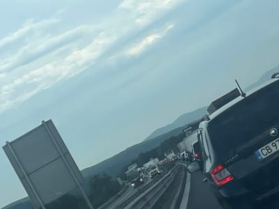 The image depicts a traffic jam on a highway near Vakarel, Bulgaria. The sky is overcast with scattered clouds. In the foreground, a black Skoda car is visible, with its license plate reading "CB 97 XX". Further ahead, several trucks and cars are stopped on the highway. The road appears to be a multi-lane highway with concrete barriers. In the background, rolling hills covered in trees are visible, suggesting a rural or semi-rural setting. The overall impression is one of congestion and slow-moving traffic.