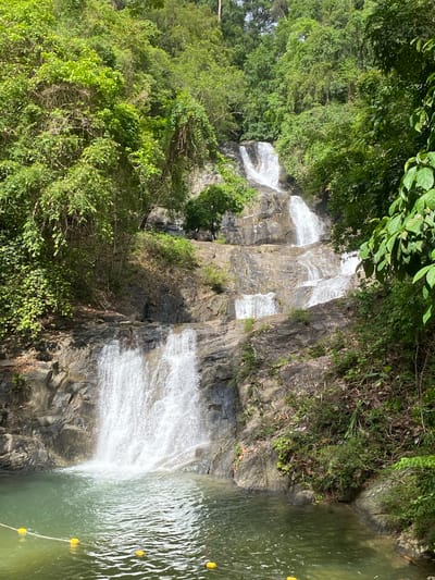 The image depicts a tiered waterfall cascading down a rocky cliff face into a pool of water, surrounded by lush green vegetation. The location is identified as Tambon Thai Mueang, Thailand. The water flows down multiple levels, creating a series of small falls. The pool at the bottom is lined with a rope and yellow buoys. The surrounding area is densely covered with trees and plants, growing on both sides of the waterfall.