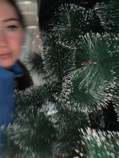 The media file is a close-up, blurry, vertical photograph featuring a person's face in the foreground and the branches of a Christmas tree in the background. The person's face is partially obscured, with only their left eye and forehead visible, suggesting they are looking towards the camera. Their expression is not clearly discernible due to the blur. They appear to be wearing a blue garment.
The Christmas tree branches are dark green and appear to be dusted with artificial snow. The branches are densely packed and fill the majority of the frame. Some red and brown elements, possibly pinecones or ornaments, are visible deep within the branches. The lighting is low, creating dark shadows and a festive, indoor atmosphere, likely within a home. The blurriness of the image suggests movement during the capture, possibly from the person or the camera.
No visible text is present in the image. The context of Старые Туймазы, Russia, suggests this photo was taken during the winter holiday season. The overall impression is an intimate, candid moment captured during preparations or celebrations for Christmas.