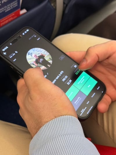 A close-up, slightly overhead image captures an individual holding a black smartphone with both hands, positioned within an aircraft cabin. The smartphone displays an active application with a dark interface. Centered on the screen is a circular profile image depicting a person in a dark t-shirt and head covering, making a peace sign gesture against an outdoor backdrop. Visible text below the profile includes "hopper rank is #79," "392.03," and "$2.2K." The right side of the screen features green panels with "$233.4" and options such as "Transfer Funds" and "Withdraw ATM," indicative of a financial or cryptocurrency-related application. The right thumb is positioned over the screen for interaction. The left wrist is covered by a light blue, ribbed sleeve. The background consists of blurred dark blue airplane seats and a partially visible red and white safety card with Russian text ("РОССИИ") inserted into a seat-back pocket. A light beige object, possibly a headrest or cushion, is also discernible. The scene is located within an airplane at or en route to Yemelyanovo International Airport (Емельяново (аэропорт)), Russia.