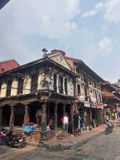 The image depicts a street scene in Lalitpur, Nepal, dominated by an old, multi-story building with traditional architecture. The building features ornately carved wooden pillars and frames, and its facade is adorned with numerous electrical wires. Several people are standing in front of the building, some appearing to be engaging in commerce or observation. A blue tarp is covering merchandise on the ground near the base of the building. Two scooters are parked in front of the building. The street is paved with uneven stone and appears well-worn. Signage and advertising are visible on the upper floors of the building. The sky is cloudy and overcast.