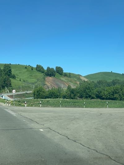 In the city of Altaiskoe, Russia, a clear blue sky stretches over a rolling green landscape. In the foreground, a paved road with visible lane markings and some dark, wet patches suggests recent precipitation or road maintenance. The road curves slightly, leading the eye towards a grassy embankment.

Along the edge of the road, a series of black and white striped bollards are spaced out, marking the boundary of the road or a potential hazard. Further back, a white guardrail runs along a raised section of the road that appears to be a bridge or overpass. A circular traffic sign with a red border and a horizontal bar indicates a prohibition, possibly "no entry" or "closed."

The middle ground is dominated by a verdant hillside, dotted with various trees. The hillside exhibits a change in terrain, with a section appearing barren and showing layers of earth in shades of brown and reddish-brown, suggesting erosion or a cut in the landscape. This section is partially covered by sparse vegetation.

Beyond this, more trees and lush green vegetation create a dense line, separating the immediate hillside from the distant landscape. The background features rolling, green hills that recede into the distance, their shapes softened by the atmospheric perspective. The overall impression is a rural or semi-rural setting, likely a roadside view in a hilly, possibly mountainous region. No people are visible in the image.