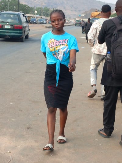 A young woman walks toward the viewer on an asphalt road. She wears a light blue t-shirt tied at the waist, a black pencil skirt with a red stain, and patterned sandals. A green minivan is parked on the left. Behind her, other people are walking in the same direction, along with a truck with people on top of the cargo area. A hazy mountain range is visible in the background.
