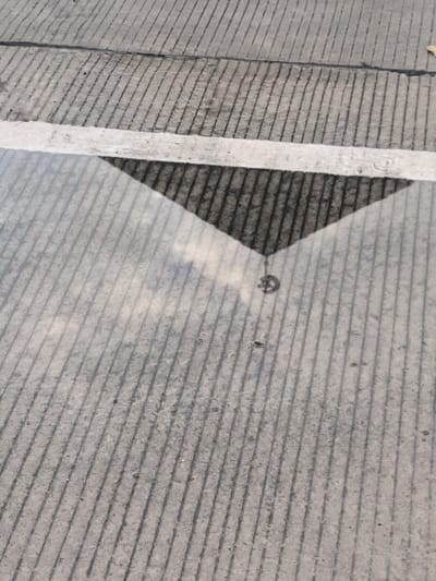 The image presents a ground-level view of a concrete surface, likely a road or paved area in Keureuweungkrueng, Indonesia. The surface is characterized by numerous parallel grooves running diagonally across the frame, providing texture and linearity. A prominent white line, painted horizontally, bisects the image roughly through its middle.
Below this white line, a puddle of water has formed, creating a reflective surface that covers the lower half of the photograph. Within this puddle, a dark, triangular shape is clearly reflected, resembling a shadow or a structural element above. The apex of this dark triangle points downwards into the puddle. The grooved texture of the concrete is also visible through the water's reflection, appearing lighter and somewhat blurred compared to the dry sections.
A notable detail within the reflection, near the pointed end of the dark triangle, is a small, faint circular symbol. This symbol appears to be a stylized letter 'G' or a simple logo, adding an intriguing element to the reflected scene.
The presence of the puddle indicates recent rainfall or wet conditions. The lighting suggests it is daytime. The scene is static, focusing on the interplay of concrete textures, painted lines, and reflections on a wet surface. No people or specific events are depicted.