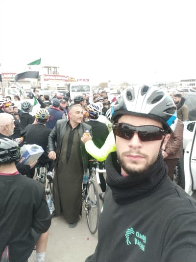 A selfie shows a man in cycling gear including a helmet, sunglasses, and neck gaiter. He wears a black sweatshirt with Arabic text. Behind him are several cyclists and pedestrians, many also wearing helmets. A man in a leather jacket and long, brown robe stands out. Bicycles are visible amongst the crowd. In the background, buildings and parked cars are seen with a flag displaying green, white, and black horizontal stripes. The scene seems to be an event or gathering in an urban environment on a cloudy day.