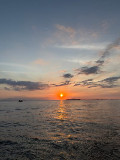 The image captures a serene sunset over the sea near Pemenang Timur, Indonesia. The sun, a brilliant orange orb, is positioned on the horizon, casting a golden glow across the water's surface. Wispy clouds, tinged with pink and orange hues, drift across a sky transitioning from pale blue to warmer tones. In the distance, a faint outline of land or mountains can be seen. A small boat is visible on the left side of the frame, sailing on the calm sea. The water itself is a deep blue, reflecting the colors of the sky and the setting sun, with gentle ripples disturbing its surface. The overall scene evokes a sense of tranquility and natural beauty.